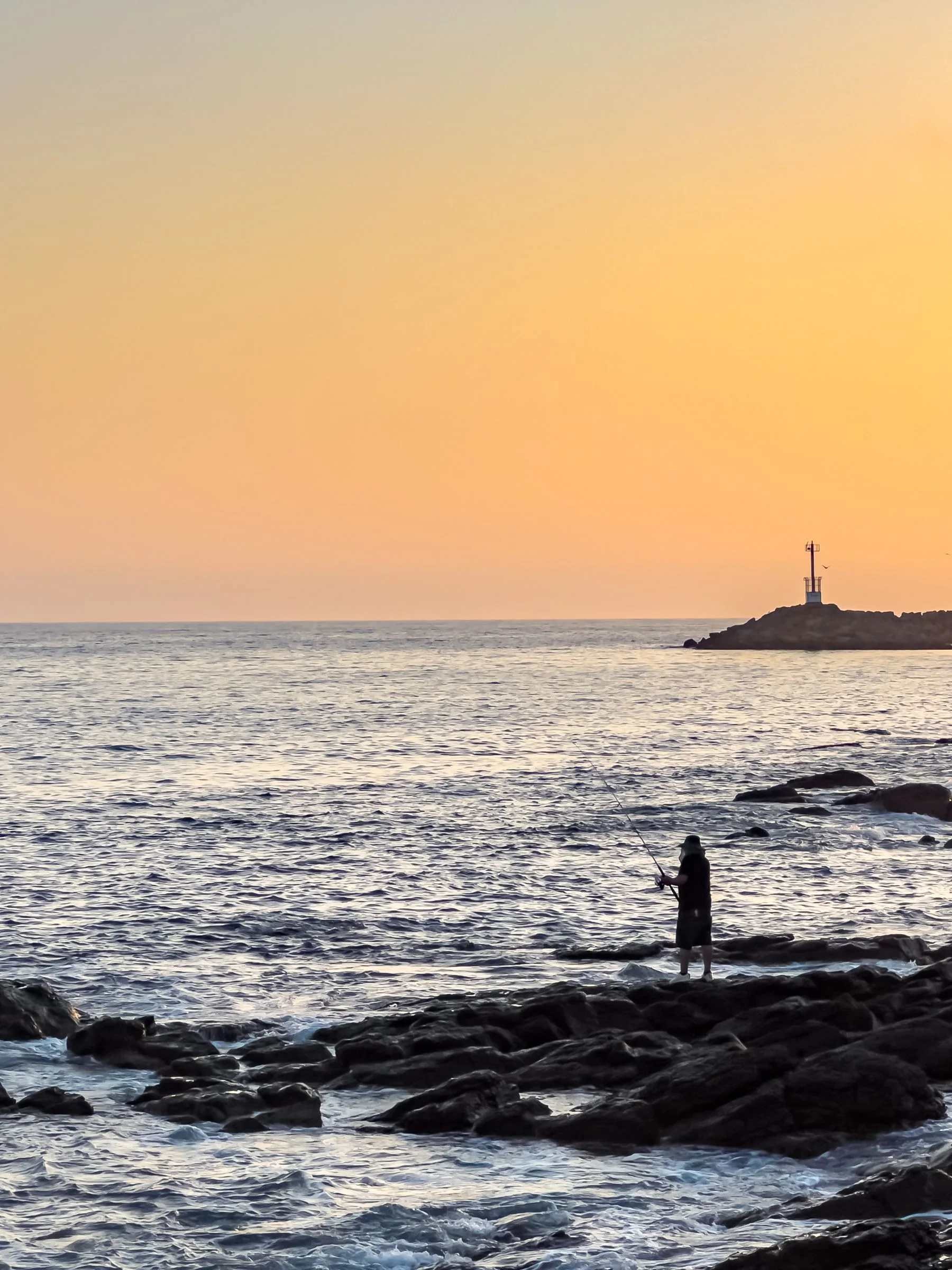 An angler on a rocky shore with a lighthouse in the distance